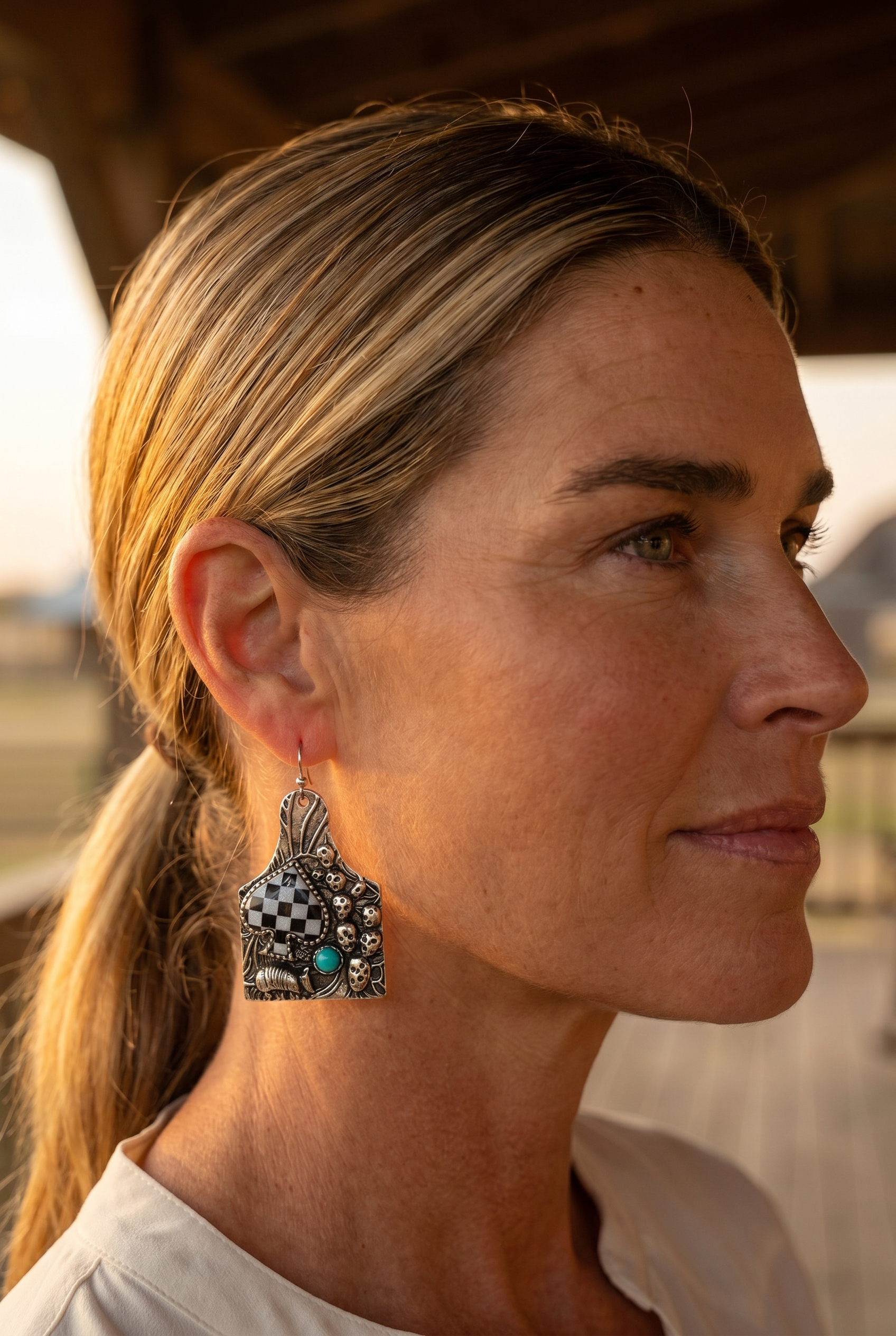 Ranch woman in golden-hour profile wearing the Cowhide & Conchos cattle-tag earrings — checkered western statement.