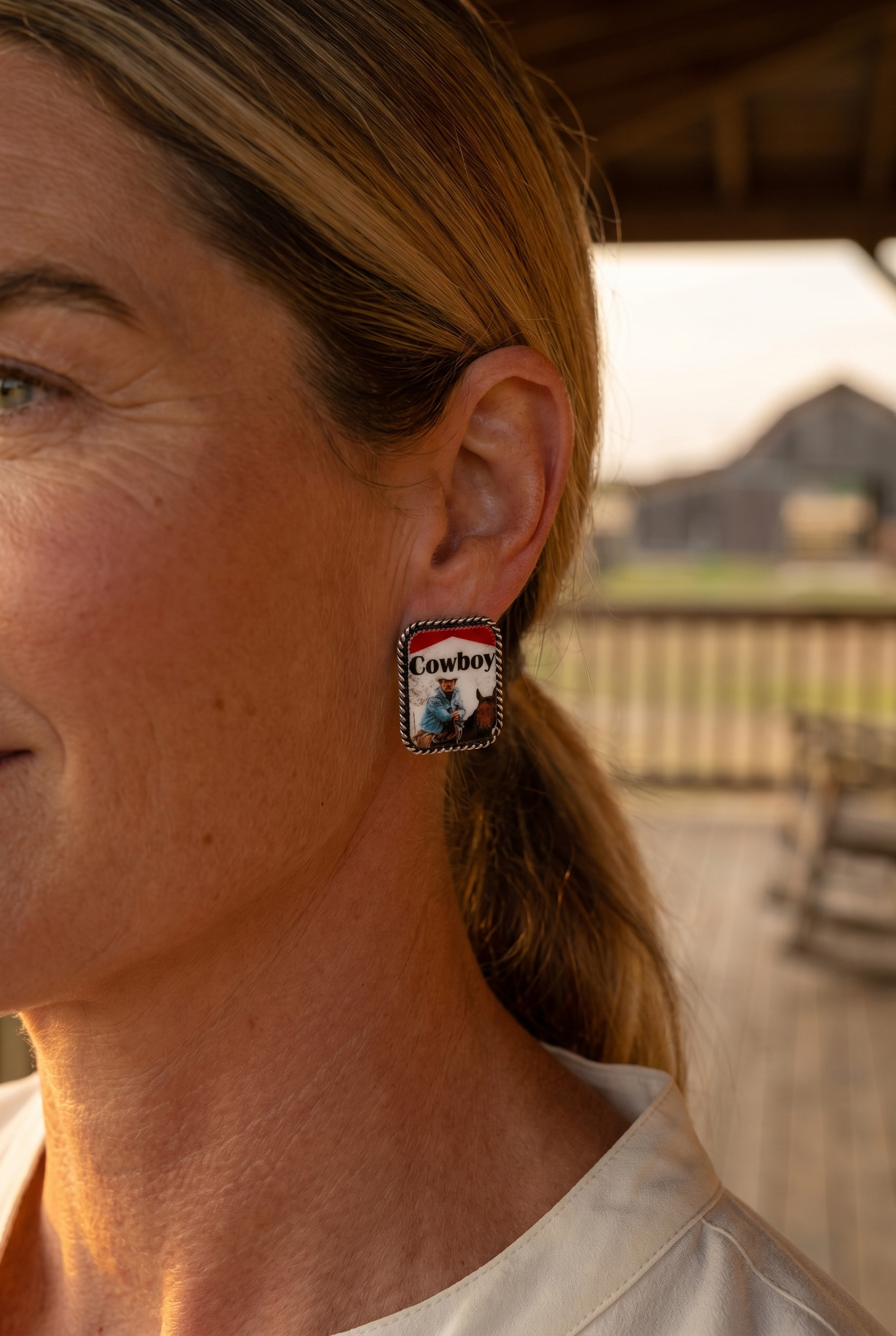 Ranch woman in golden-hour profile wearing the Cowhide & Conchos Cowboy pack earrings — playful western novelty.