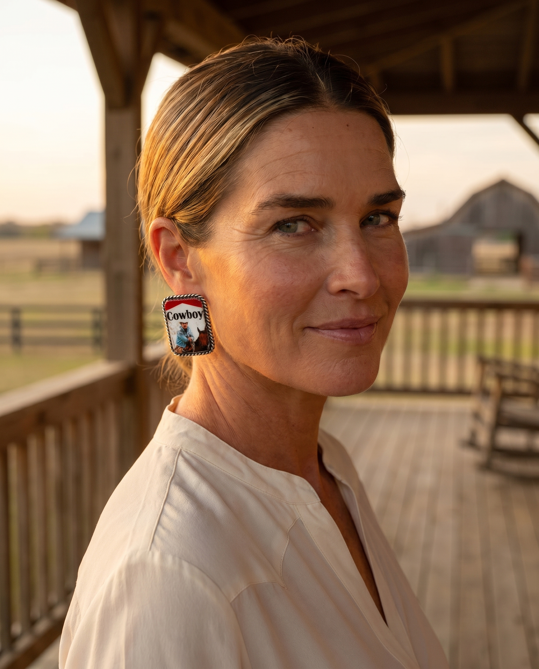 Ranch woman on a sunlit porch wearing the Cowhide & Conchos Cowboy pack earrings — western humor made everyday.