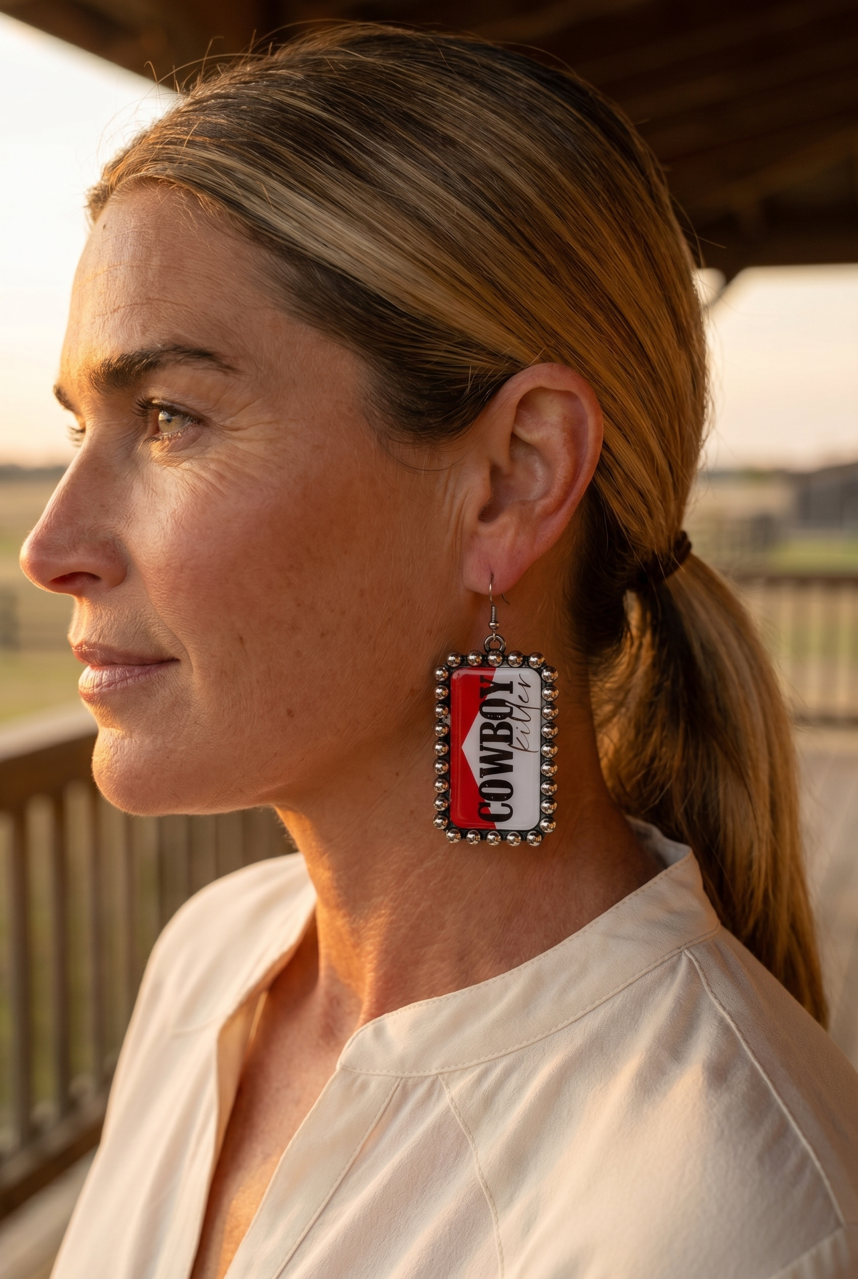 Ranch woman in golden-hour profile wearing the Cowhide & Conchos Cowboy Killer earrings — bold western novelty.