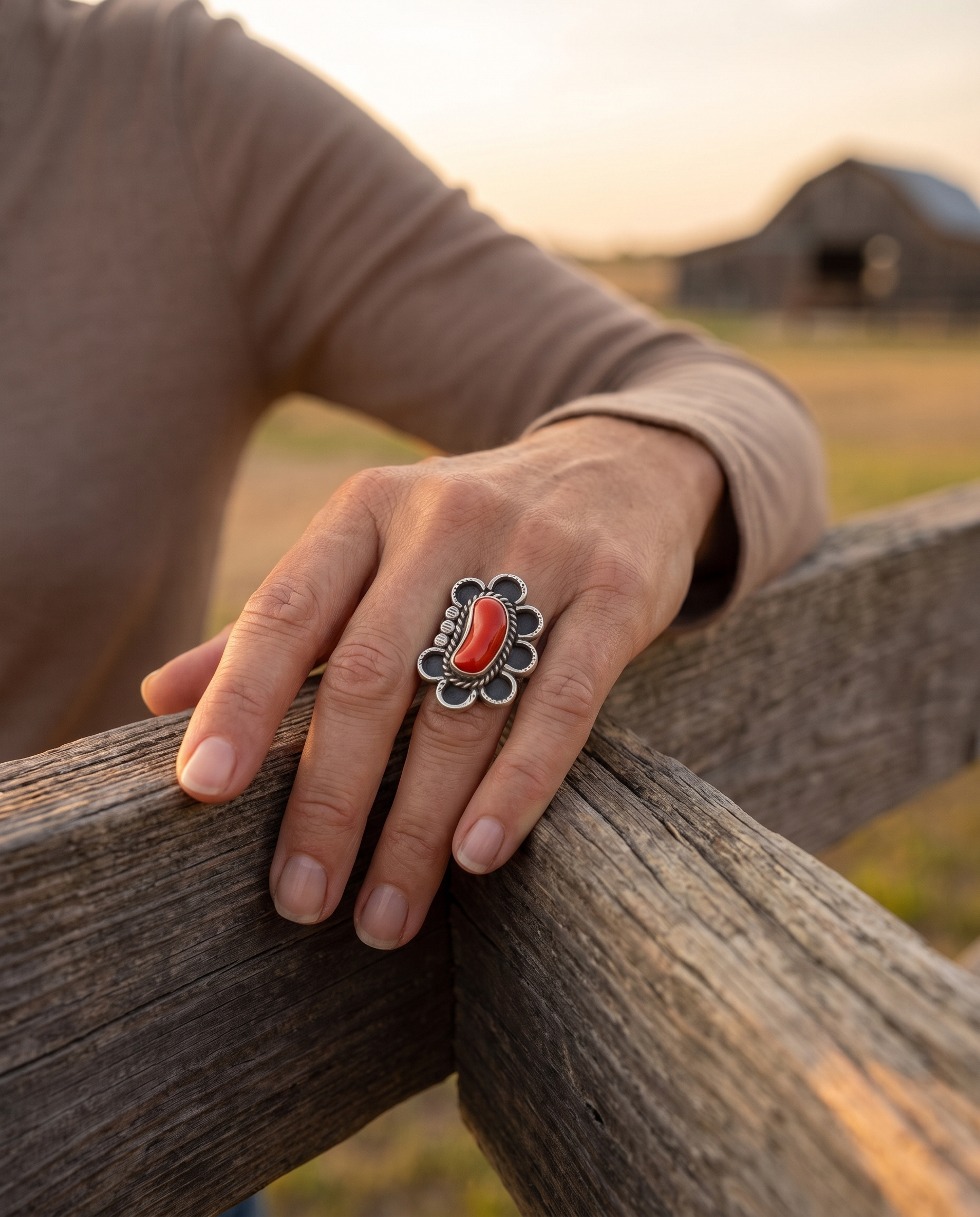 A hand on a ranch fence rail wearing the Cowhide & Conchos Annie Chapo hand-stamped sterling silver coral ring — vintage western style.