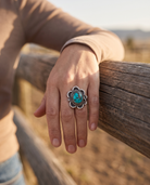A hand on a ranch fence rail wearing the Cowhide & Conchos turquoise cabochon flower-base ring — feminine western style.