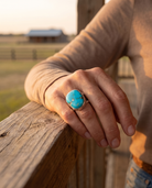 A hand on a ranch fence rail wearing the Cowhide & Conchos turquoise cabochon split-band statement ring — bold western style.