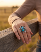 A hand on a ranch fence rail wearing the Cowhide & Conchos hand-stamped sterling silver turquoise ring — timeless western style.